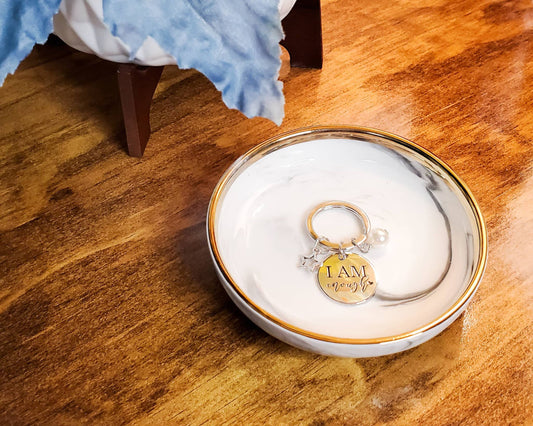 Picture of a silver keychain in a porcelain dish. The keychain says " I am Enough" on a round stainless steel circle about the size of a quarter. "I am" is written in all caps and "enough" is written in a pretty script below "I am." There are two little dangles on either side of the  I am Enough. One is a little hollow star. The other is a little white pearl-like bead.